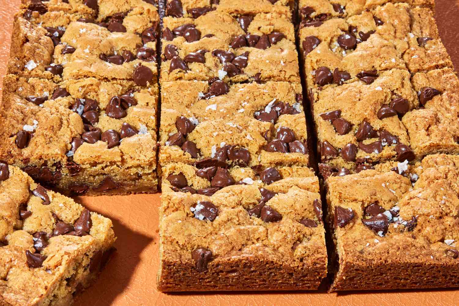A metal baking pan filled with golden blonde brownies cut into squares, with visible chocolate chips and crisp browned edges on a light kitchen counter
