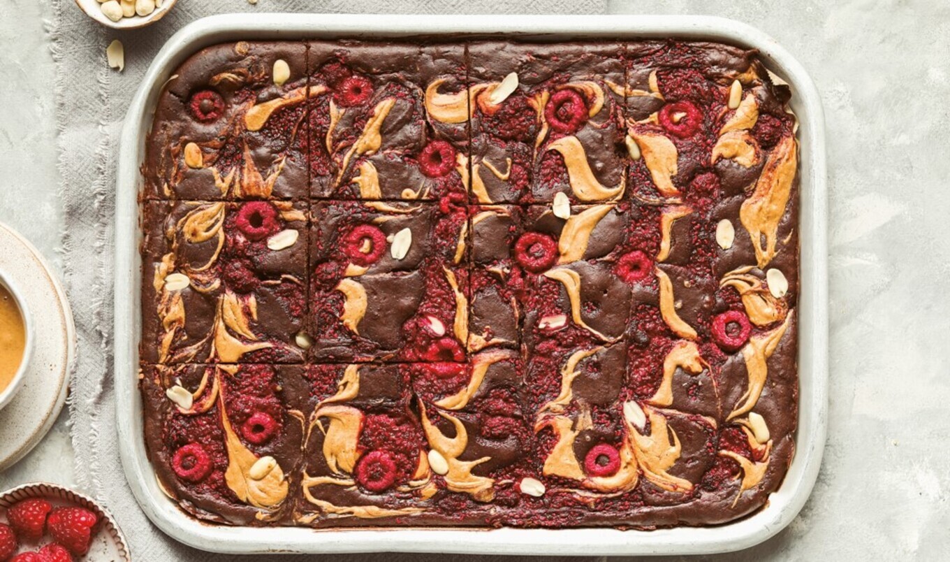 A metal baking pan filled with thick, fudgy vegan brownies with a glossy crackly top, sliced into squares on a parchment-lined tray on a warm-toned kitchen counter