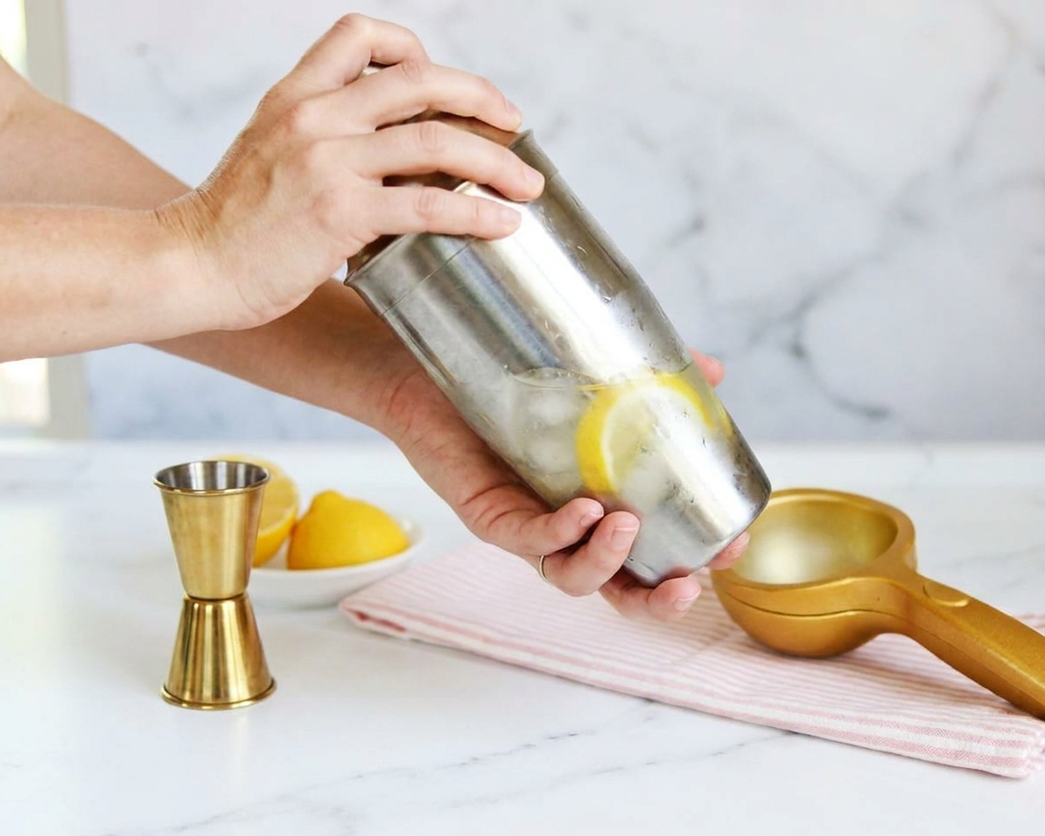 A metal cocktail shaker being shaken with lemons and a measuring jigger nearby on a clean kitchen counter
