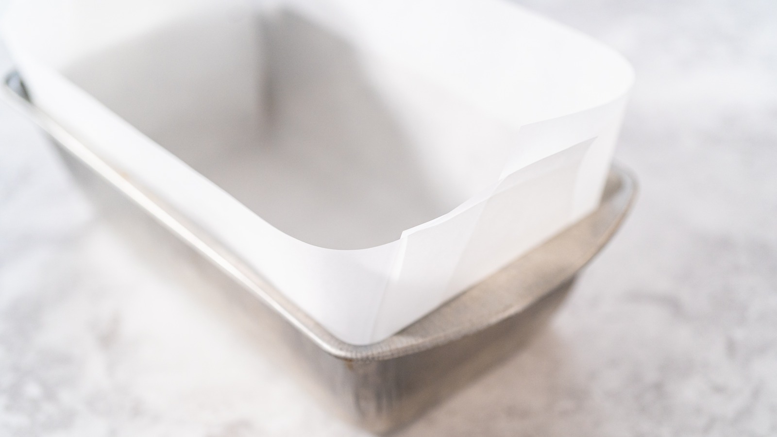 A metal loaf pan lined with parchment paper with overhang on both sides, sitting on a clean kitchen counter