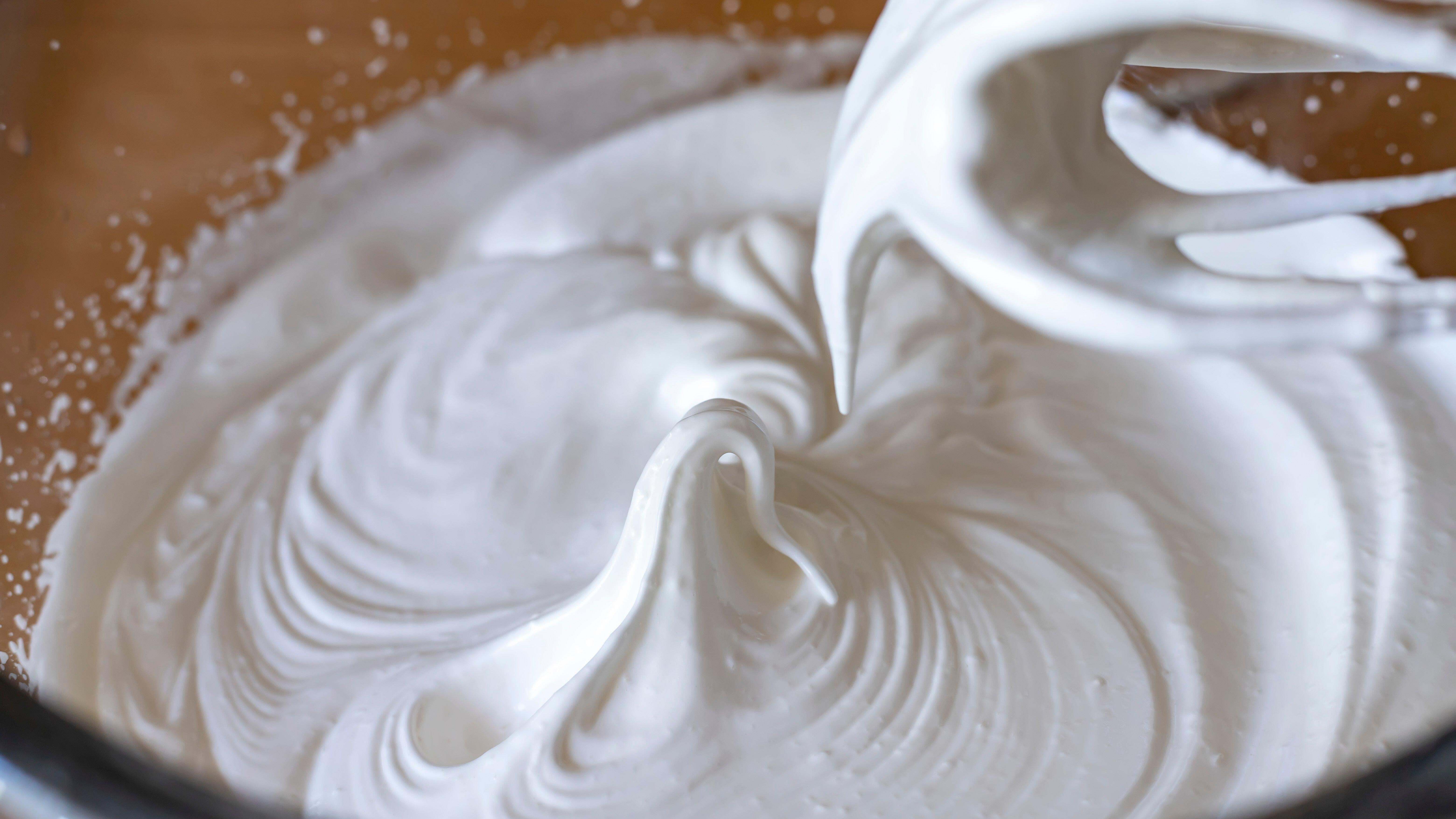 A metal mixing bowl of whipped cream at soft to medium peaks with a hand mixer resting beside it on a kitchen counter