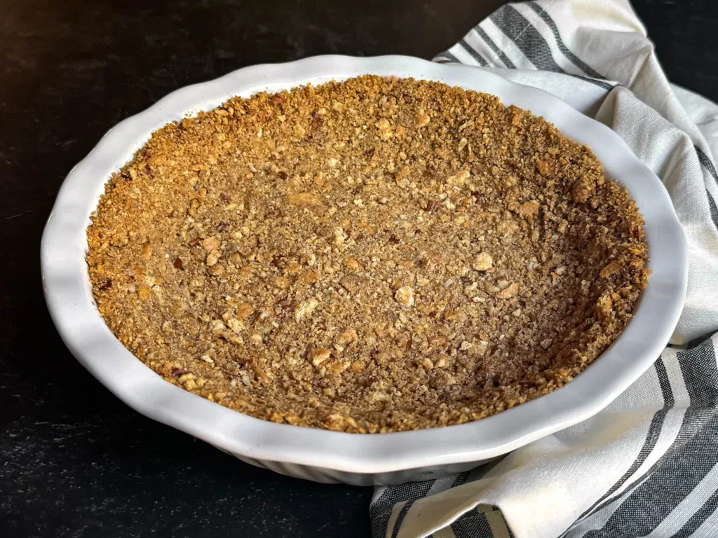 A mixing bowl filled with graham cracker crumbs, chopped toasted pecans, and melted butter being stirred into a sandy crust mixture on a kitchen counter