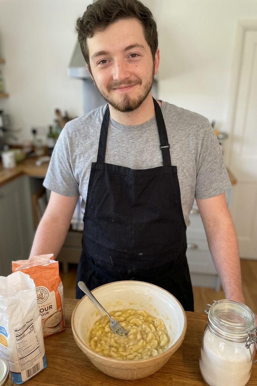 A mixing bowl filled with mashed ripe bananas and a fork resting inside, on a home kitchen counter with flour and sugar nearby, real photo style