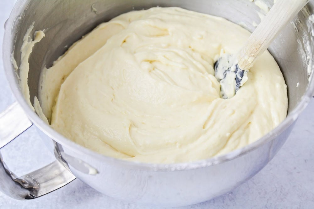 A mixing bowl filled with pale vanilla cake batter being folded with a rubber spatula on a bright kitchen counter