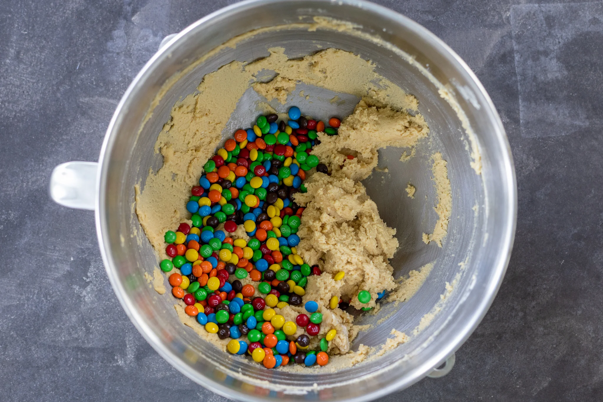 A mixing bowl of cookie dough with colorful M&Ms being folded in with a rubber spatula on a kitchen counter