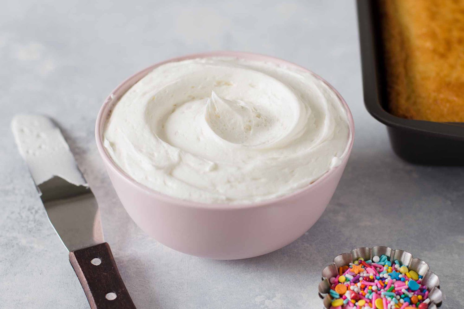 A mixing bowl of fluffy vanilla buttercream with an offset spatula resting on the rim on a bright kitchen counter