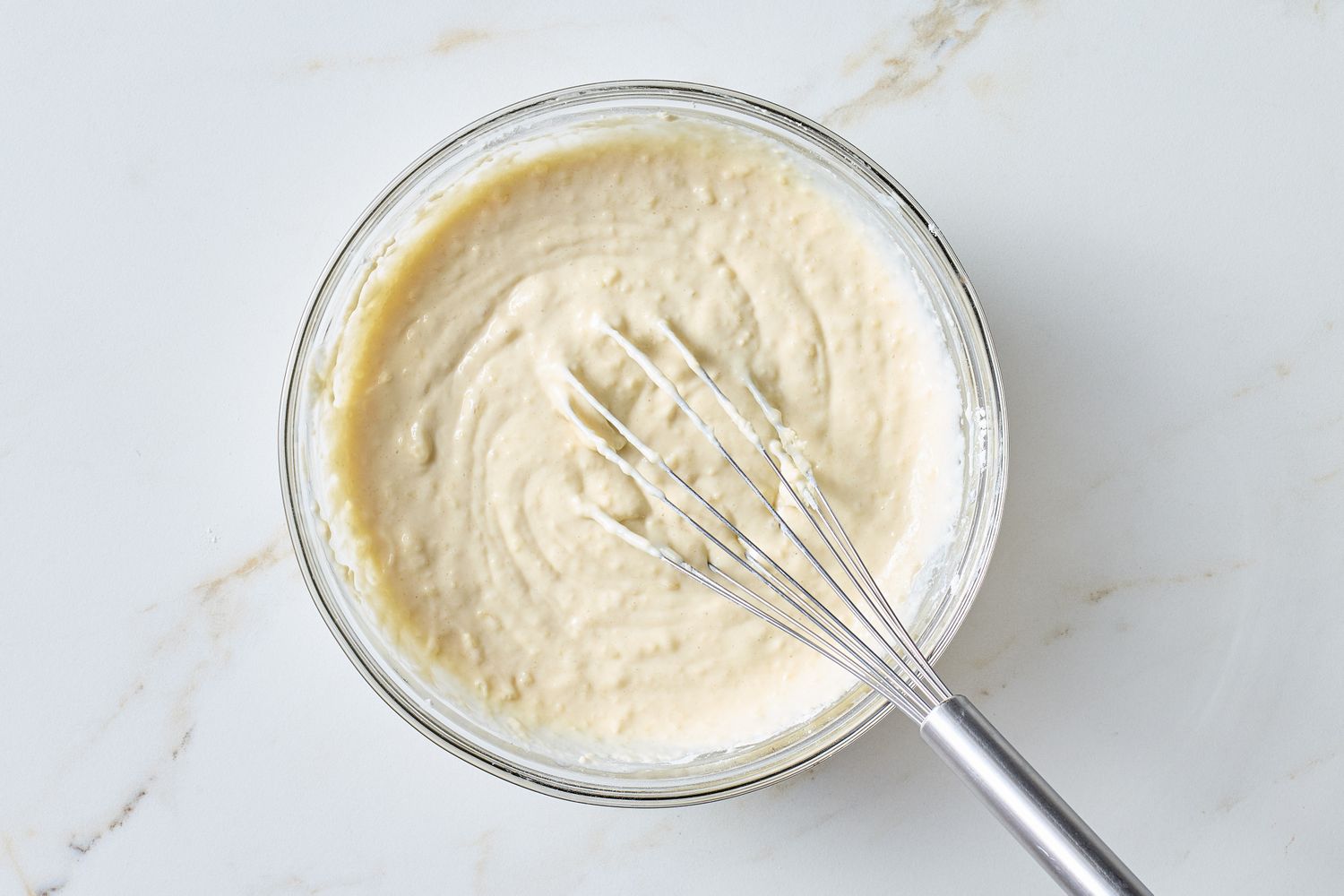 A mixing bowl of pale waffle batter with a whisk resting inside on a kitchen counter next to measuring cups