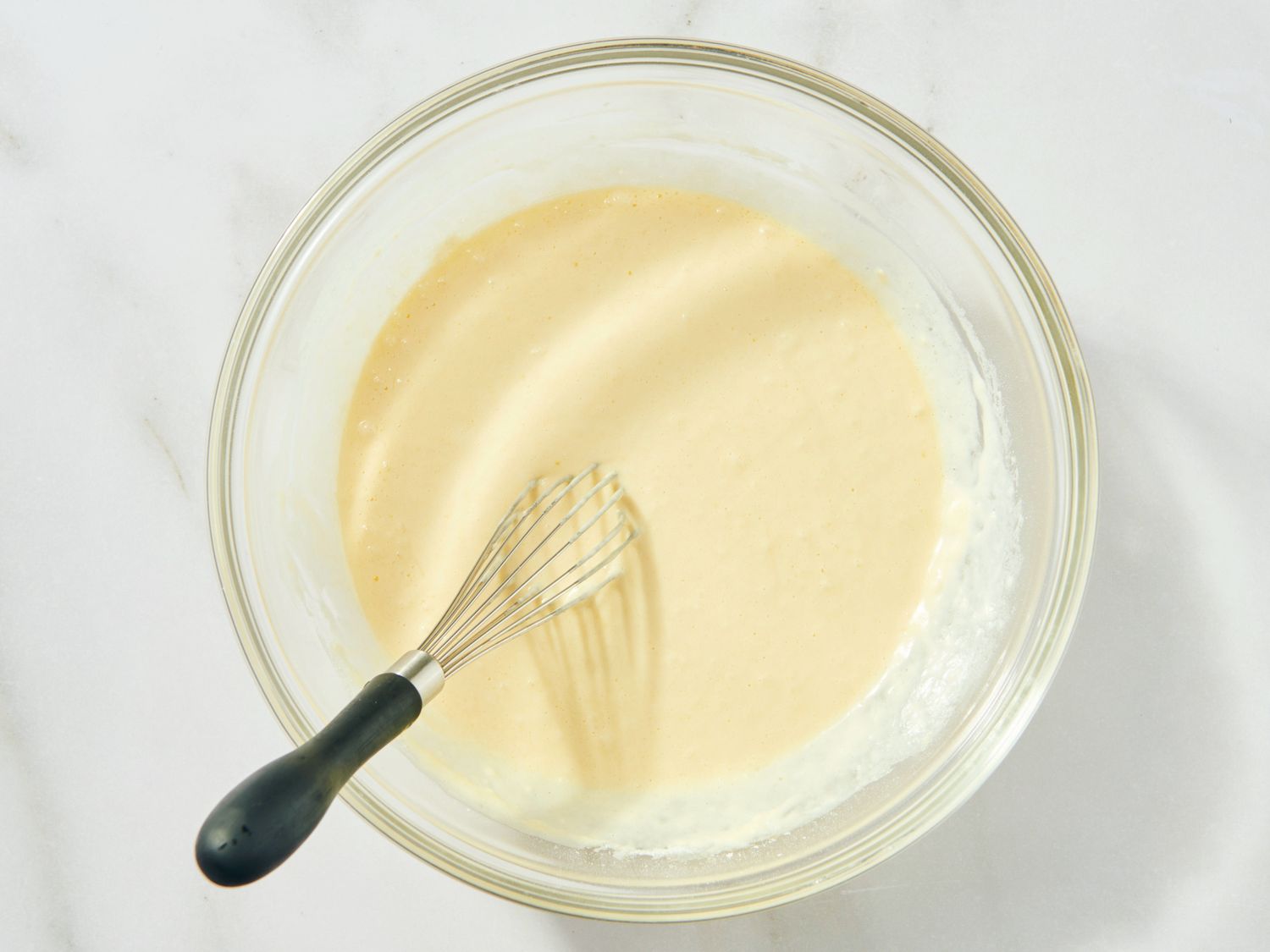 A mixing bowl of pancake batter being whisked gently with a few small lumps remaining on a bright kitchen counter