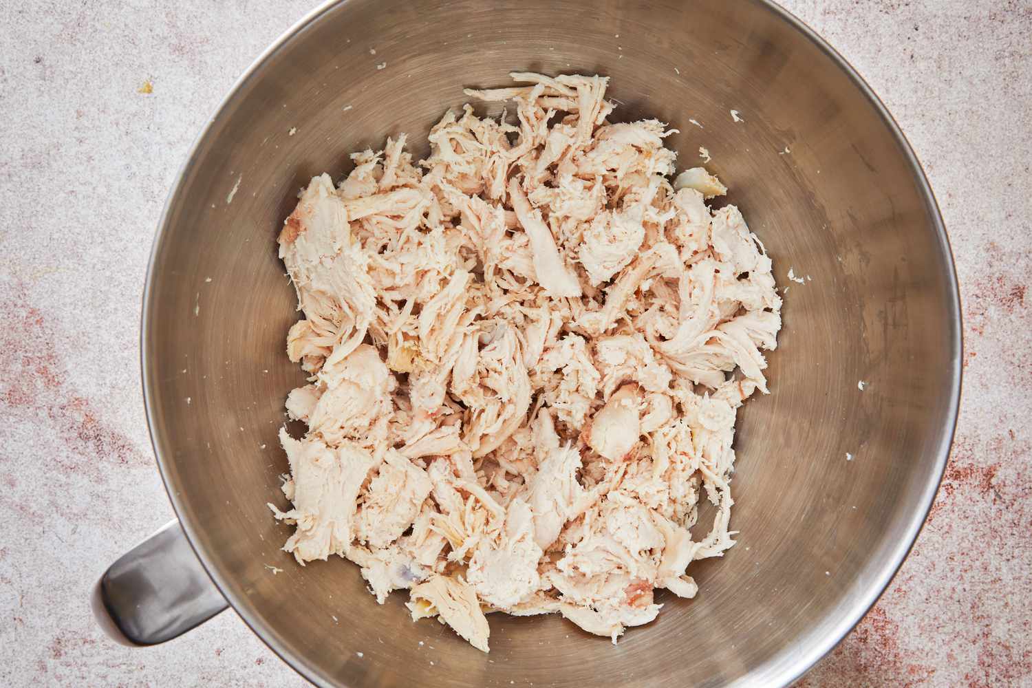 A mixing bowl on a kitchen counter with freshly shredded cooked chicken and a wooden spoon nearby, ready for making buffalo chicken dip