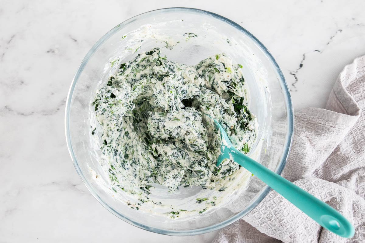 A mixing bowl on a kitchen counter with ricotta, squeezed spinach, egg, parmesan, and herbs being stirred together with a spoon