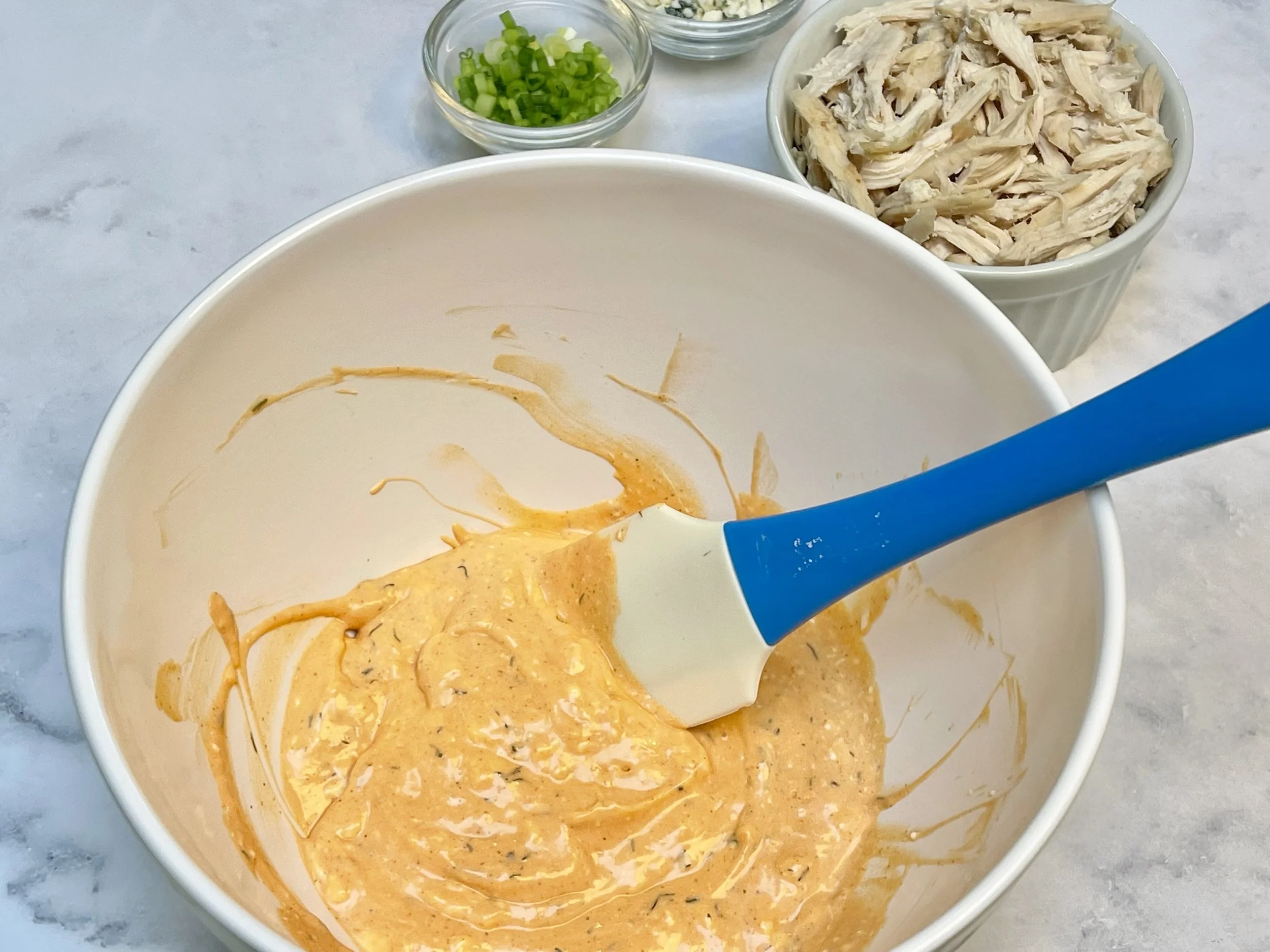 A mixing bowl on a kitchen counter with shredded chicken, softened cream cheese, buffalo sauce, ranch dressing, and shredded cheese ready to be stirred together