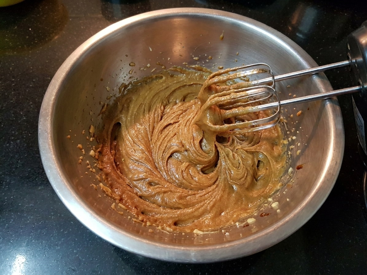 A mixing bowl with butter, brown sugar, and molasses being beaten with a hand mixer, smooth and fluffy