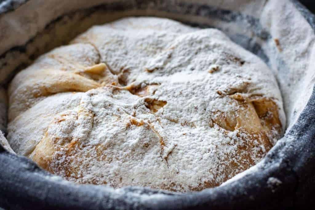 A mixing bowl with enriched bread dough being stirred, with flour dusted on the counter and a wooden spoon nearby in a bright home kitchen