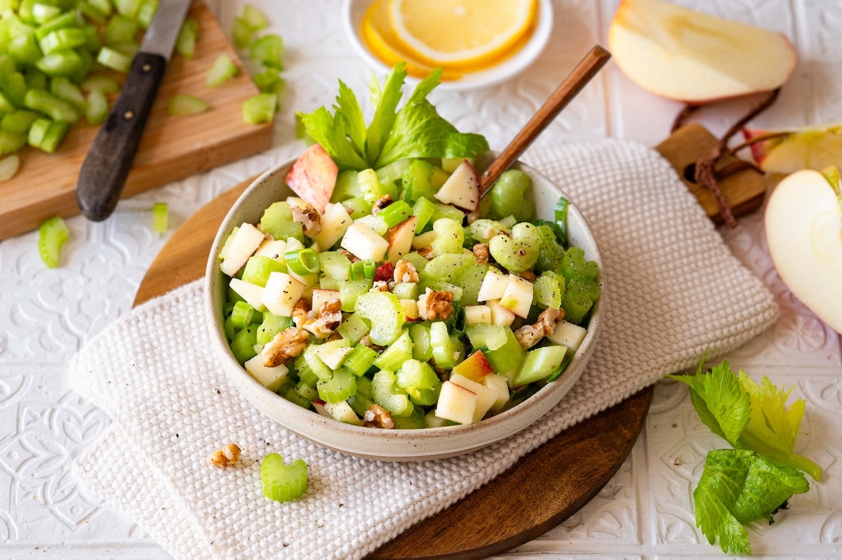 A mixing bowl with freshly chopped apple chunks and thinly sliced celery on a kitchen counter, with a chef’s knife and cutting board nearby