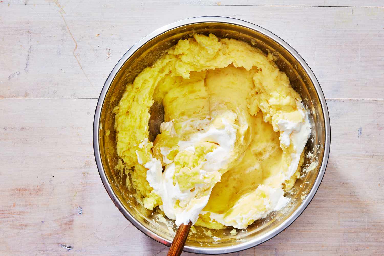 A mixing bowl with hot mashed potatoes being stirred with butter and sour cream on a kitchen counter