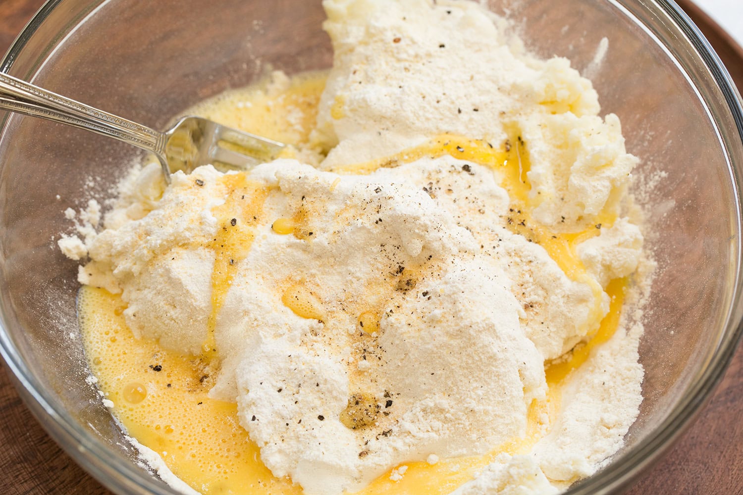 A mixing bowl with leftover mashed potatoes being stirred together with egg, flour, shredded cheese, and green onions on a home kitchen counter