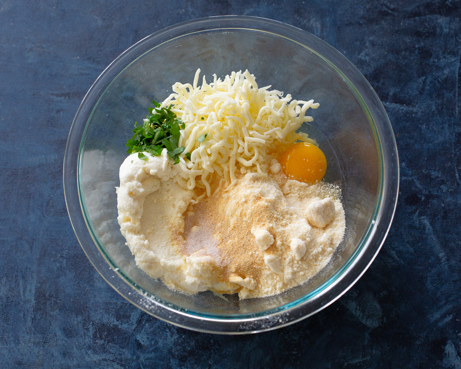 A mixing bowl with ricotta, cracked egg, grated parmesan, black pepper, and herbs ready to be stirred for lasagna filling on a kitchen counter