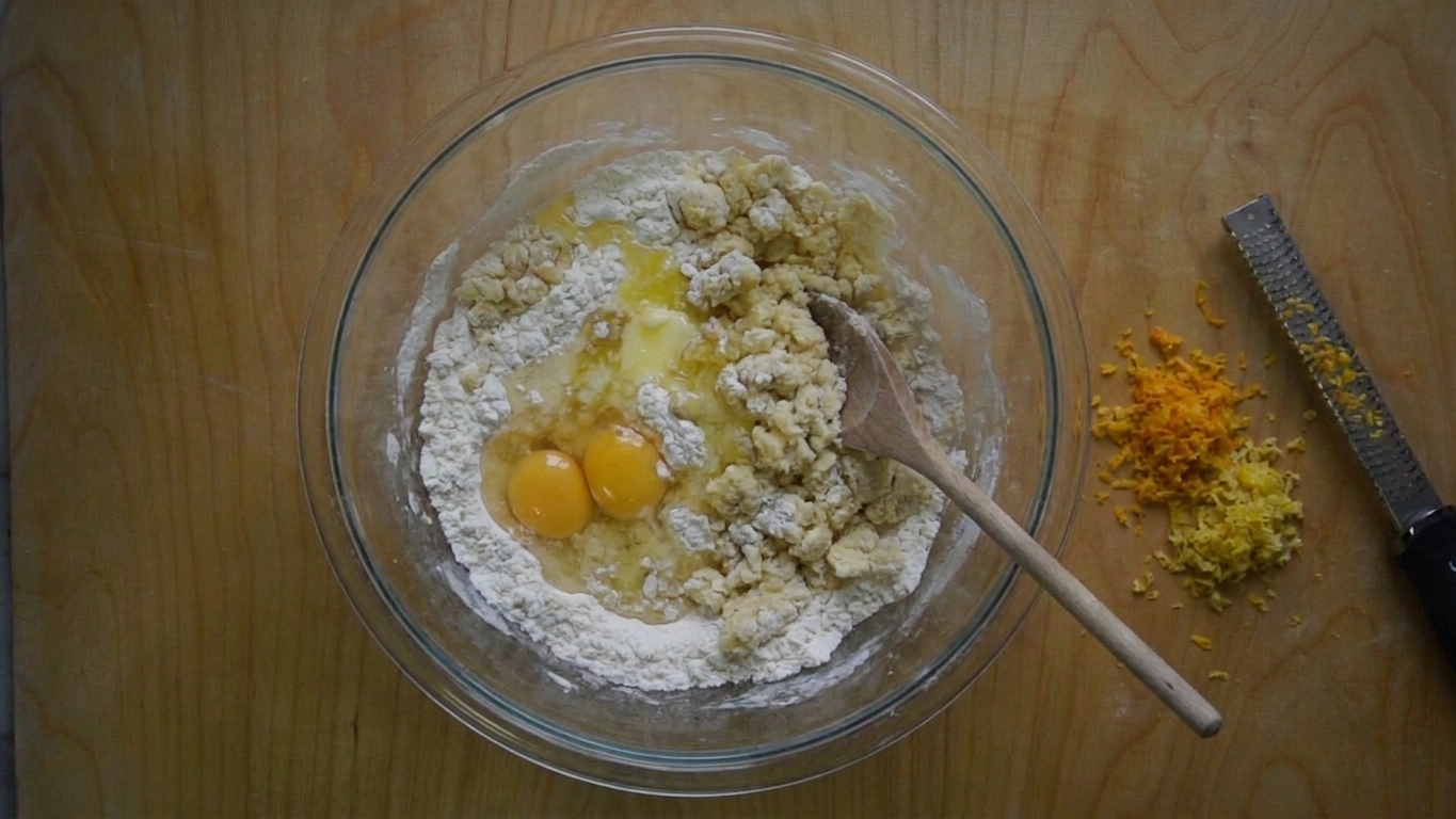 A mixing bowl with struffoli dough ingredients partially combined, with a wooden spoon and citrus zest visible on a clean kitchen counter