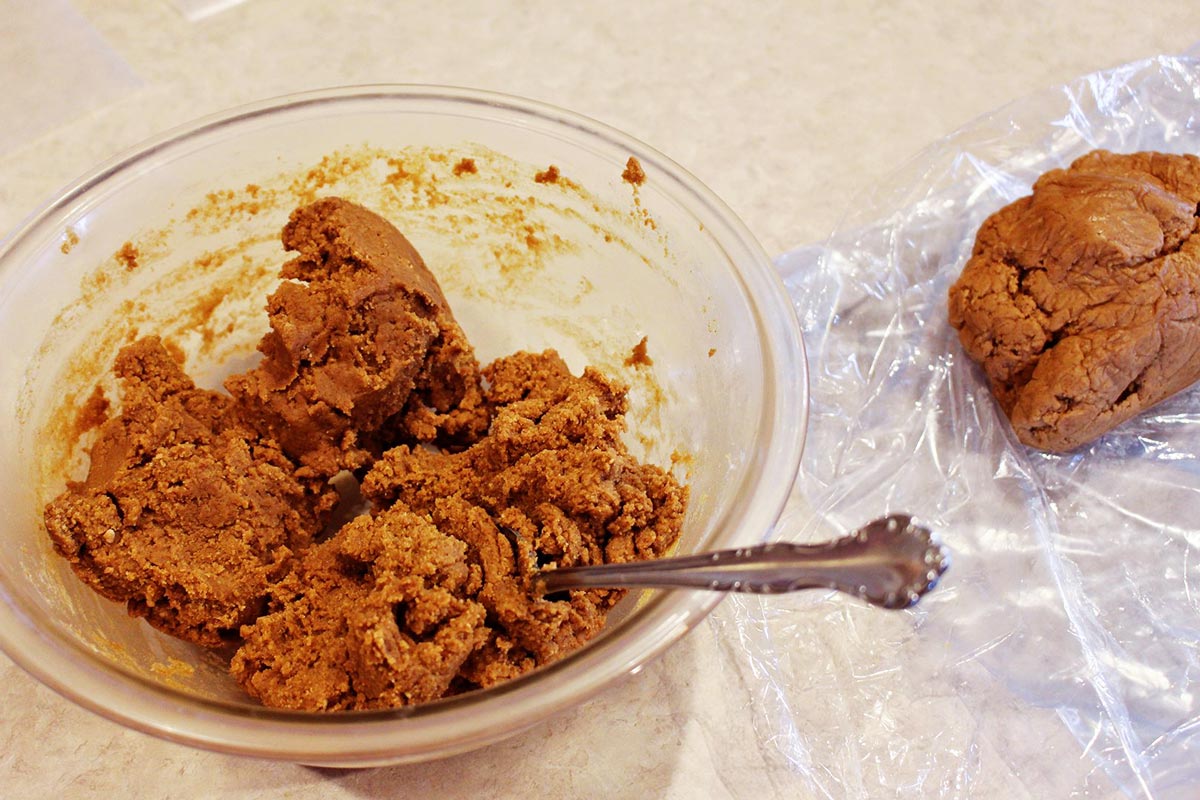 A mixing bowl with thick gingerbread cookie dough being stirred with a spatula on a kitchen counter