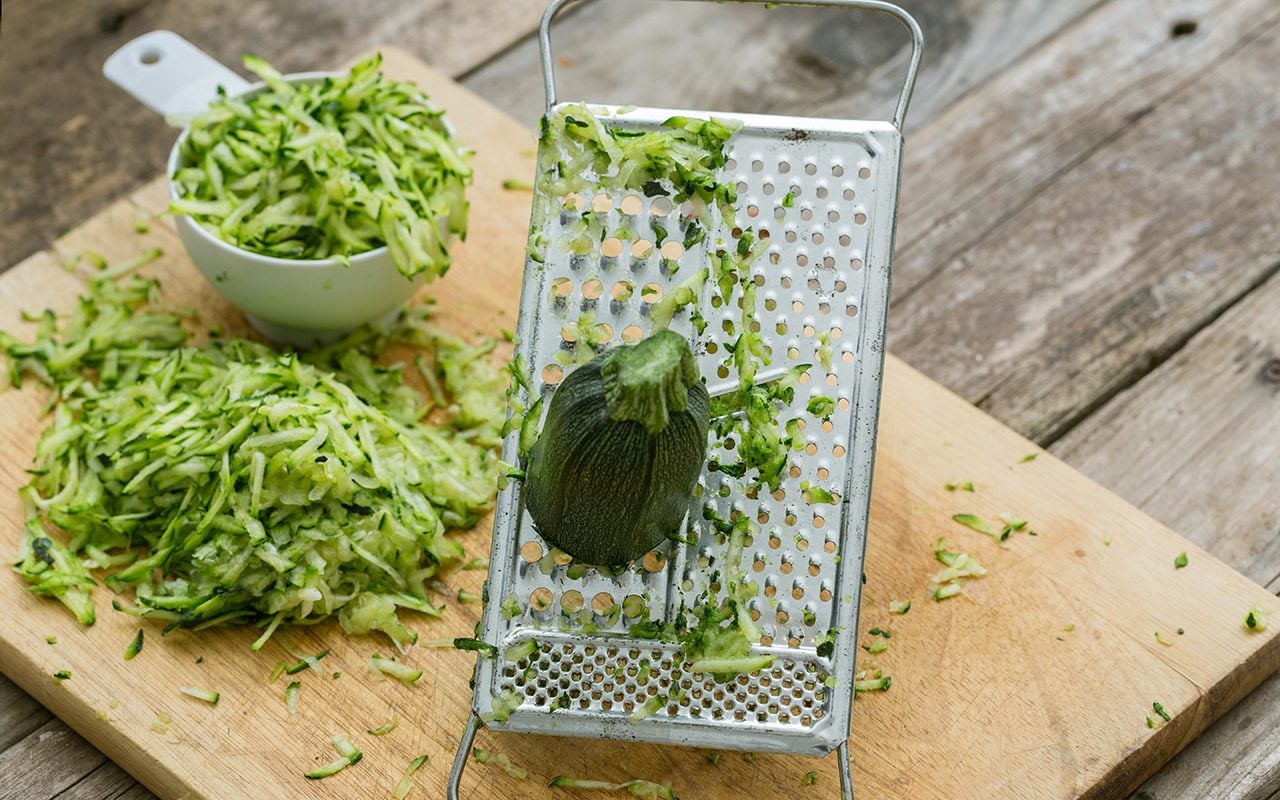 A mound of freshly shredded zucchini on a wooden cutting board beside a box grater and whole zucchini on a bright kitchen counter