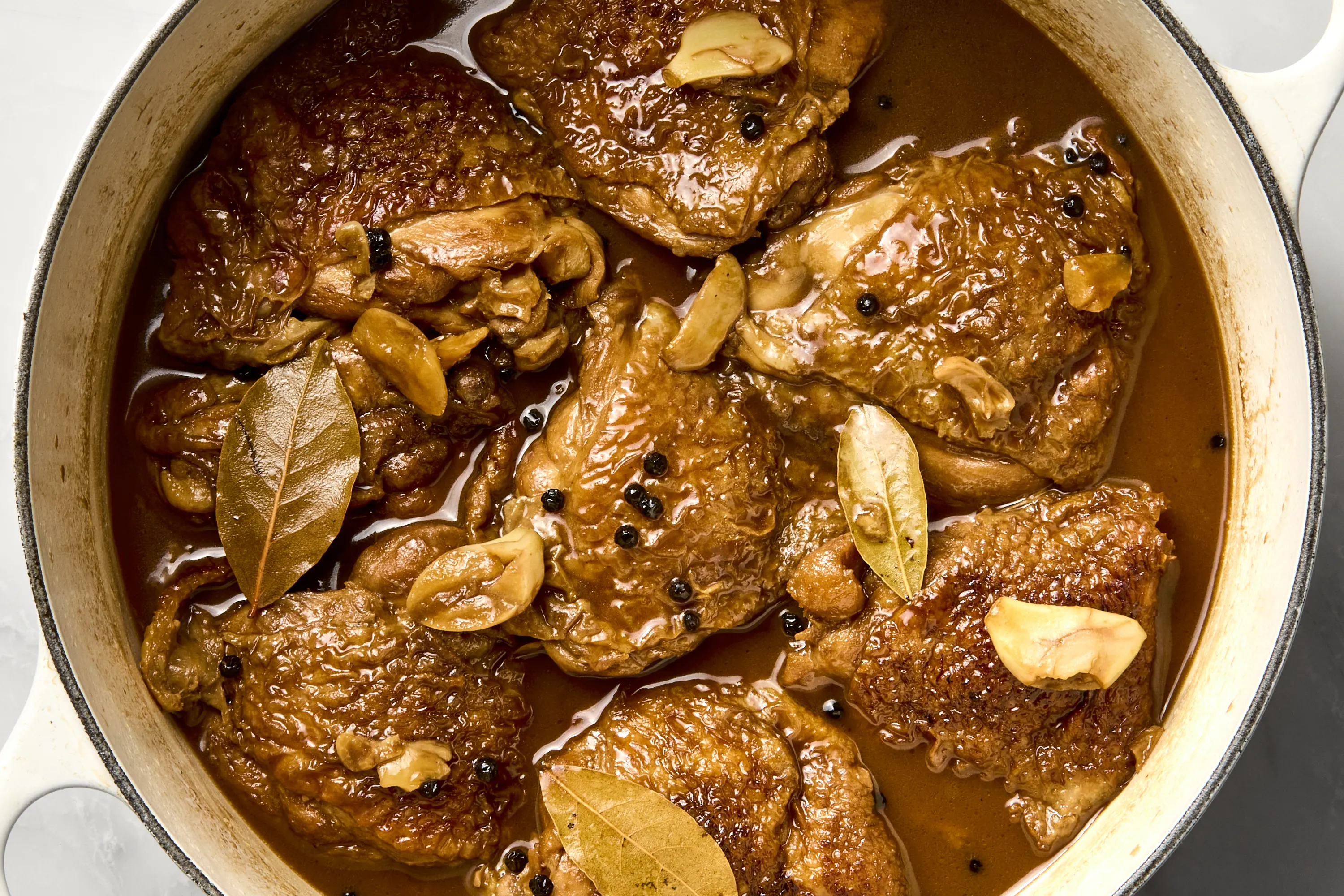 A neat arrangement of adobo ingredients on a kitchen counter: chicken thighs, garlic, soy sauce, cane vinegar, bay leaves, whole peppercorns, and a small bowl of brown sugar