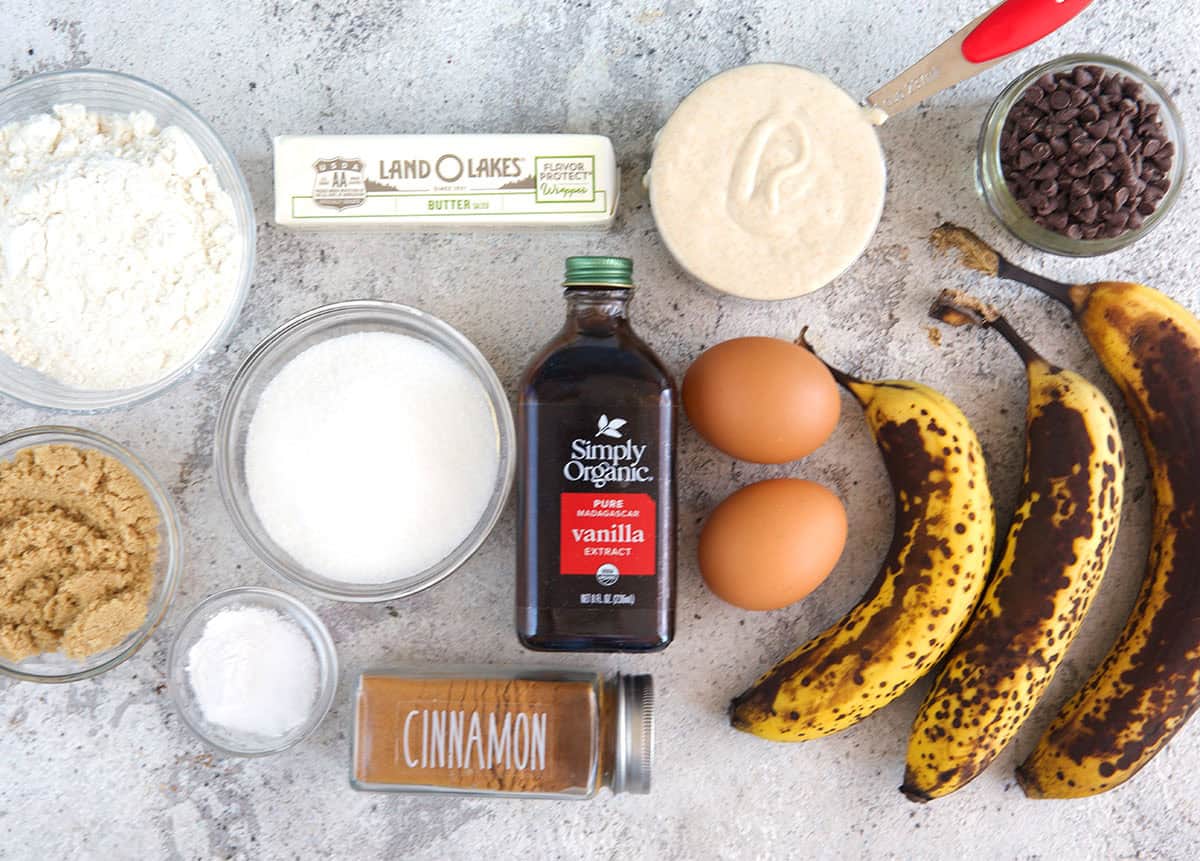 A neat arrangement of bananas, flour, sugar, sour cream, eggs, butter, vanilla, and baking soda on a light colored kitchen counter in natural window light