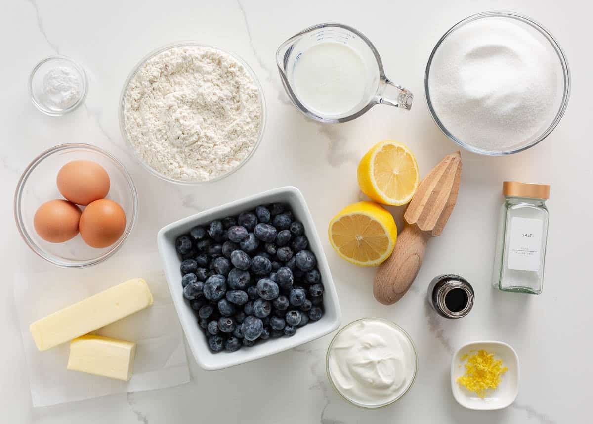 A neat arrangement of flour, sugar, butter, eggs, blueberries, lemon, and baking powder on a light stone kitchen counter with measuring cups nearby