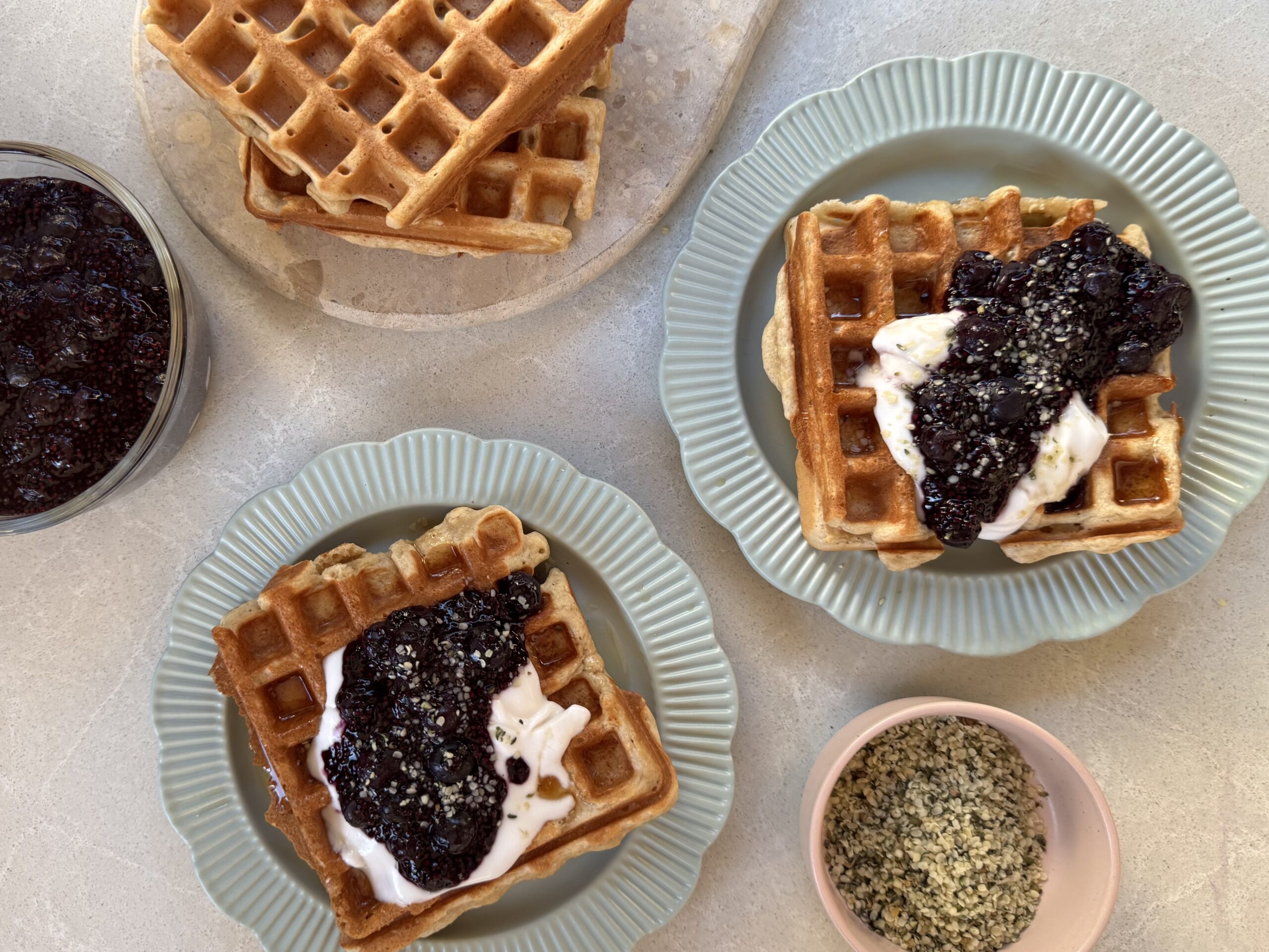 A neat arrangement of vegan waffle ingredients on a light kitchen counter, including flour, sugar, baking powder, plant milk, oil, vanilla, salt, and a small bowl of cornstarch