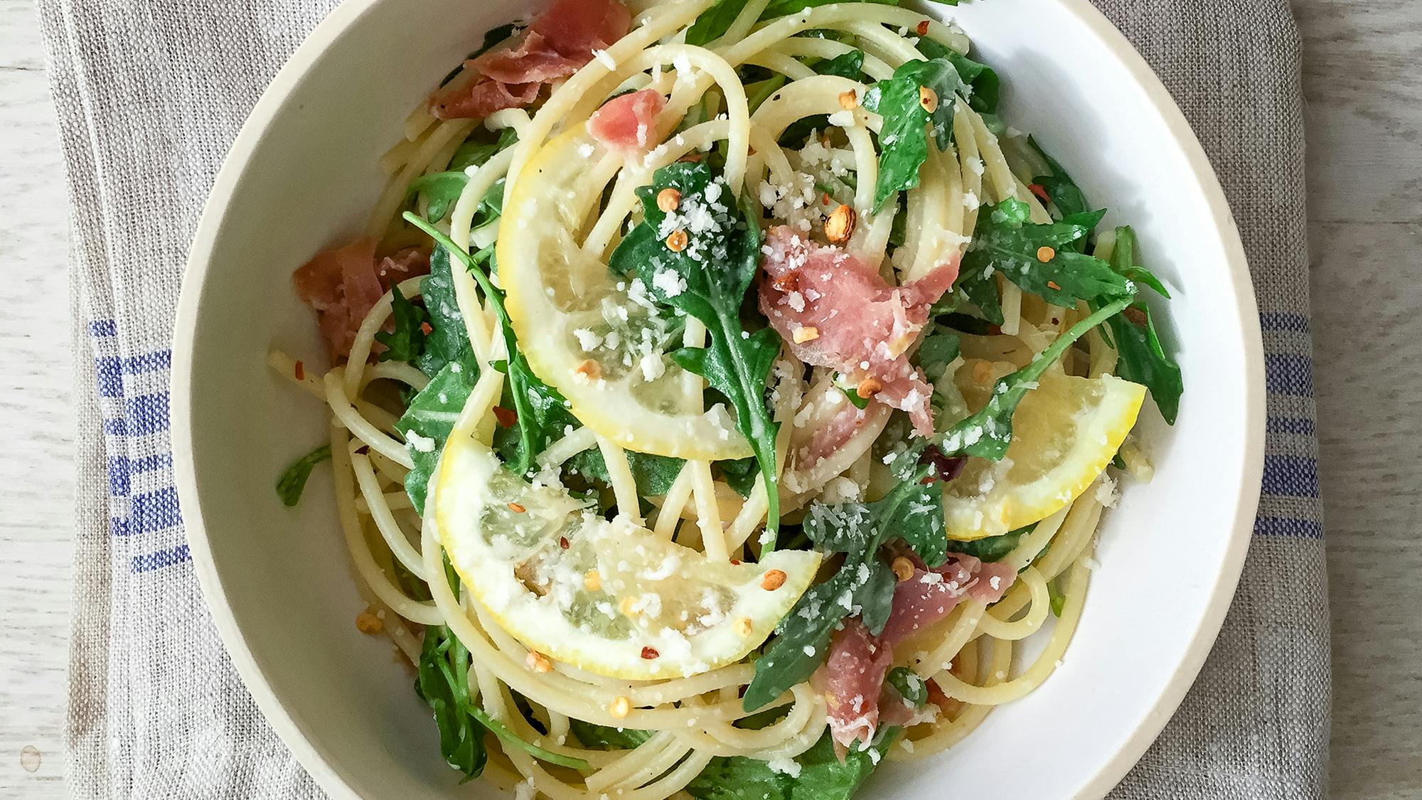 A neat ingredient setup on a kitchen counter with prosciutto, raw chicken cutlets, dry orzo, lemons, baby spinach, parmesan, garlic, and chicken broth in small bowls