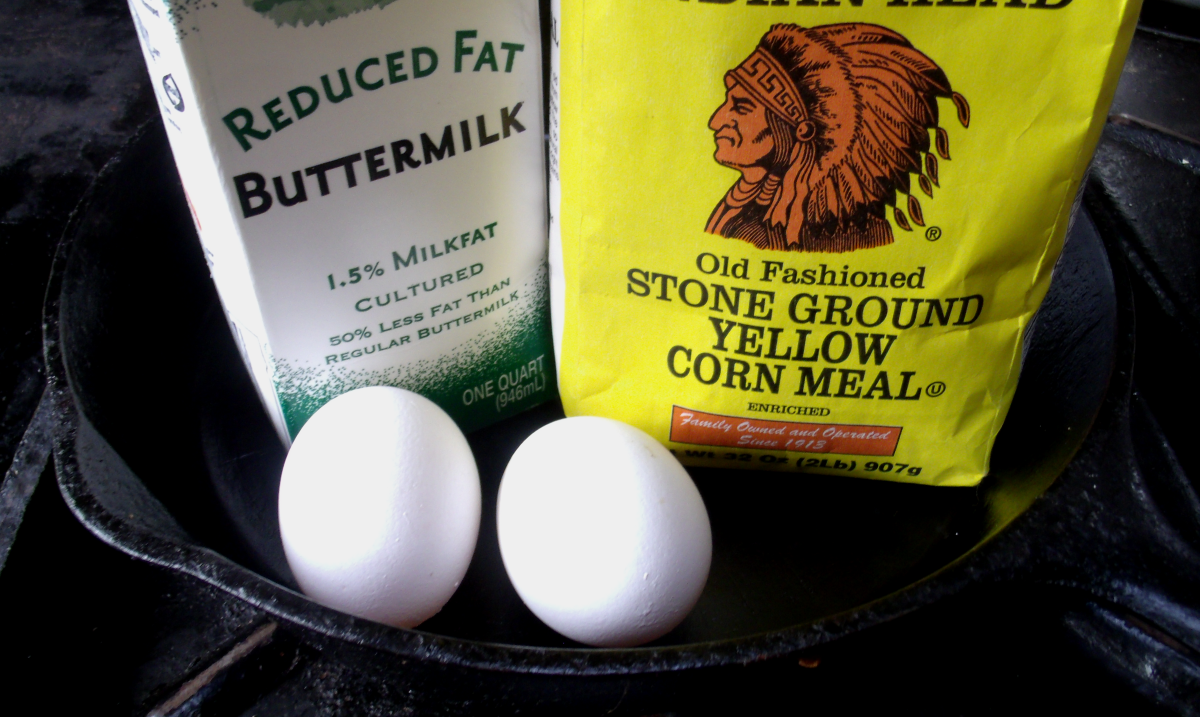 A neat lineup of cornbread ingredients on a light kitchen counter: cornmeal, flour, baking powder, baking soda, salt, eggs, buttermilk, butter, honey, and sugar