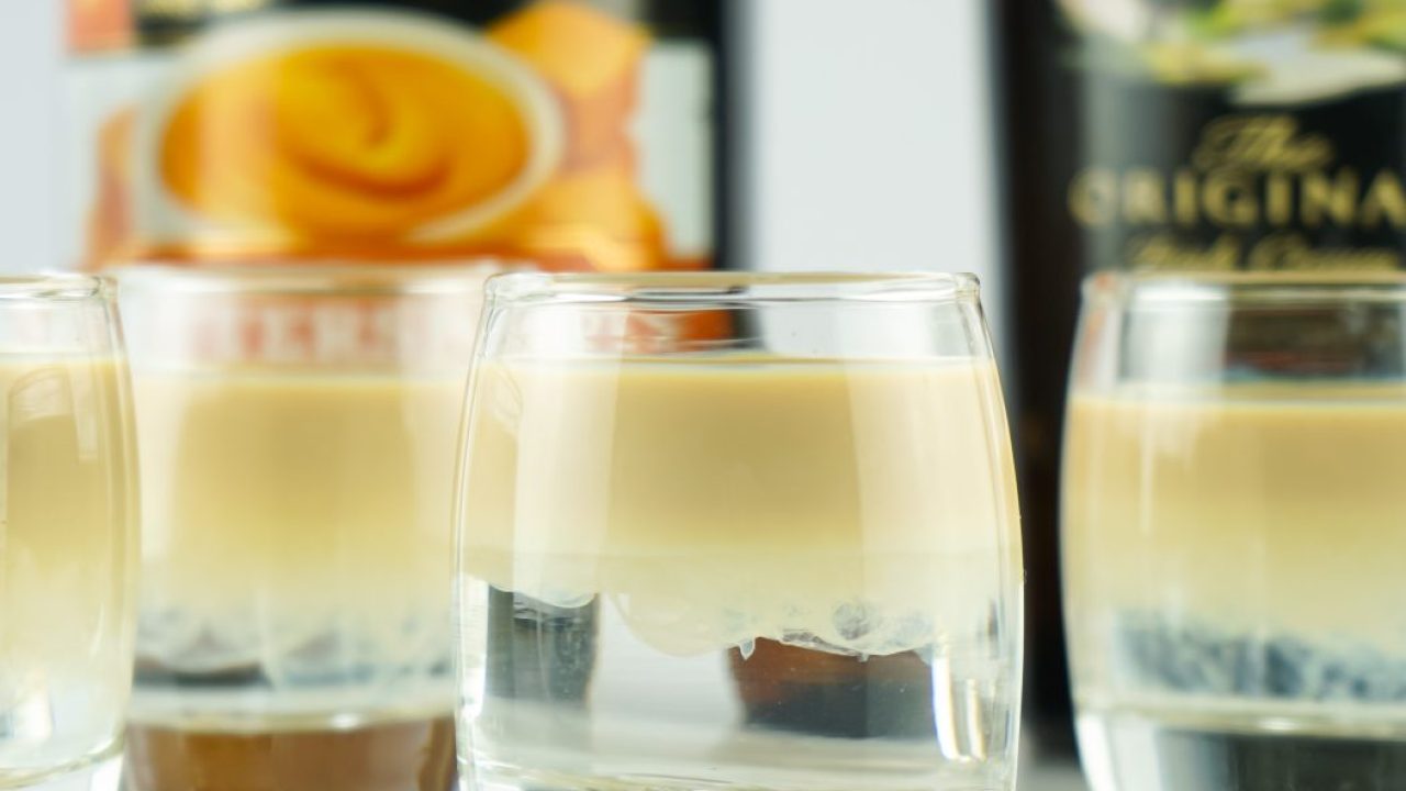 A neat row of three Buttery Nipple shots with visible layers of golden butterscotch schnapps and pale Irish cream on a warm kitchen counter under soft lighting