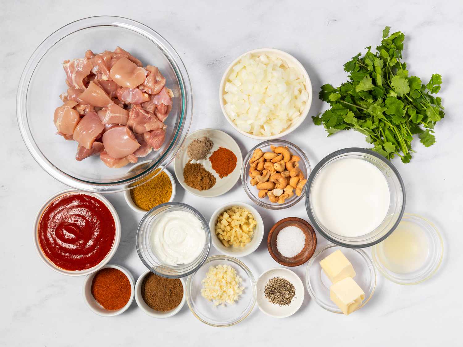 A neatly arranged set of butter chicken ingredients on a kitchen counter including chicken thighs, yogurt, canned crushed tomatoes, garlic, ginger, spices, butter, and heavy cream