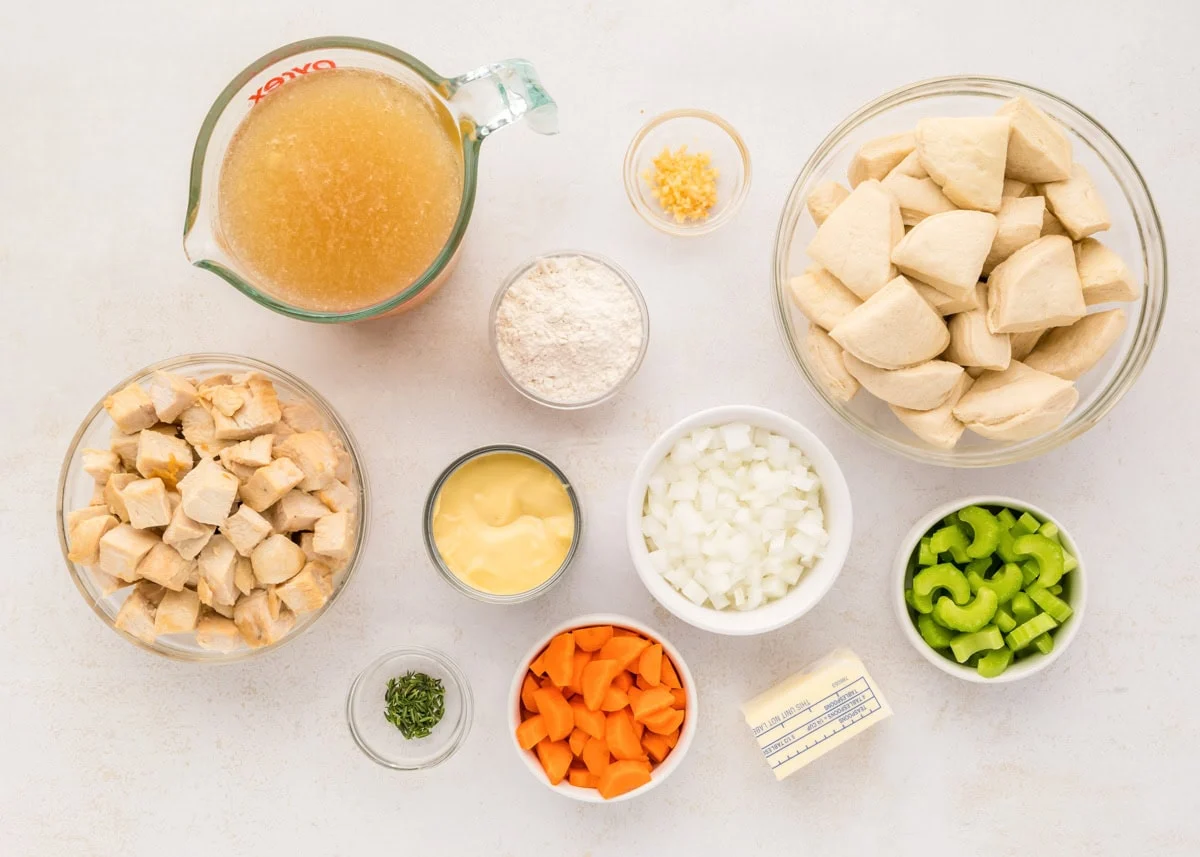 A neatly arranged spread of ingredients for chicken dumpling bake on a warm-toned kitchen counter, including cooked chicken, frozen peas and carrots, flour, milk, broth, butter, and seasonings in small bowls