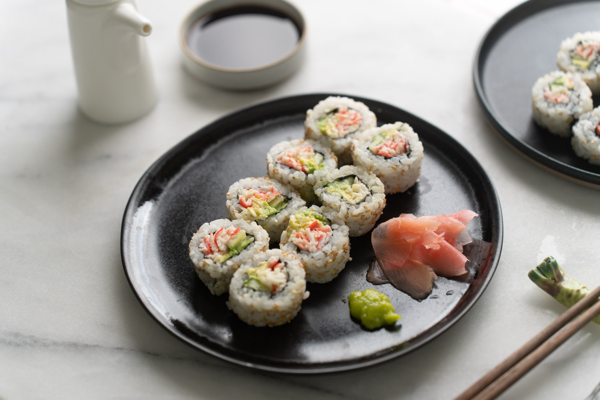 A neatly sliced California roll arranged on a ceramic plate with a small dish of soy sauce and a few slices of pickled ginger on a warm kitchen table