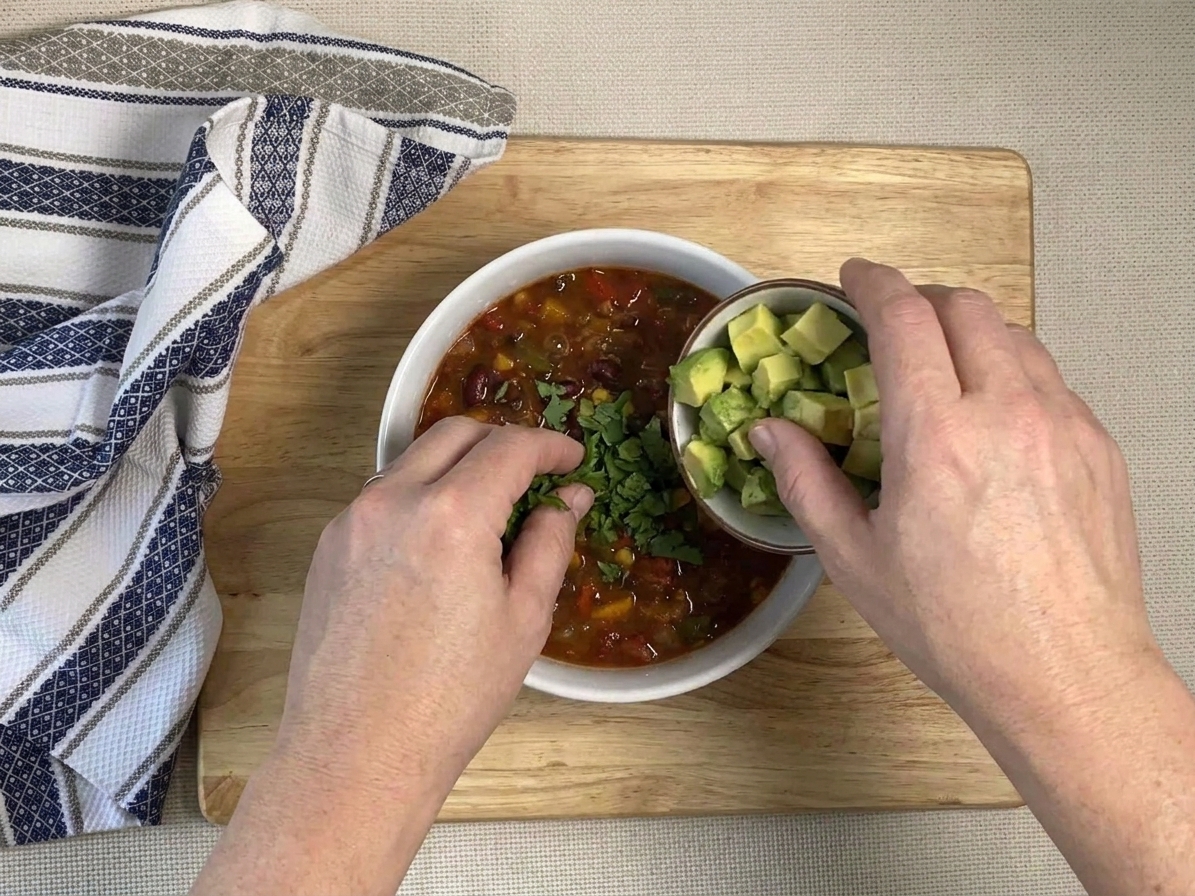 A pair of hands adding diced avocado and cilantro to a bowl of veggie chili on a kitchen counter