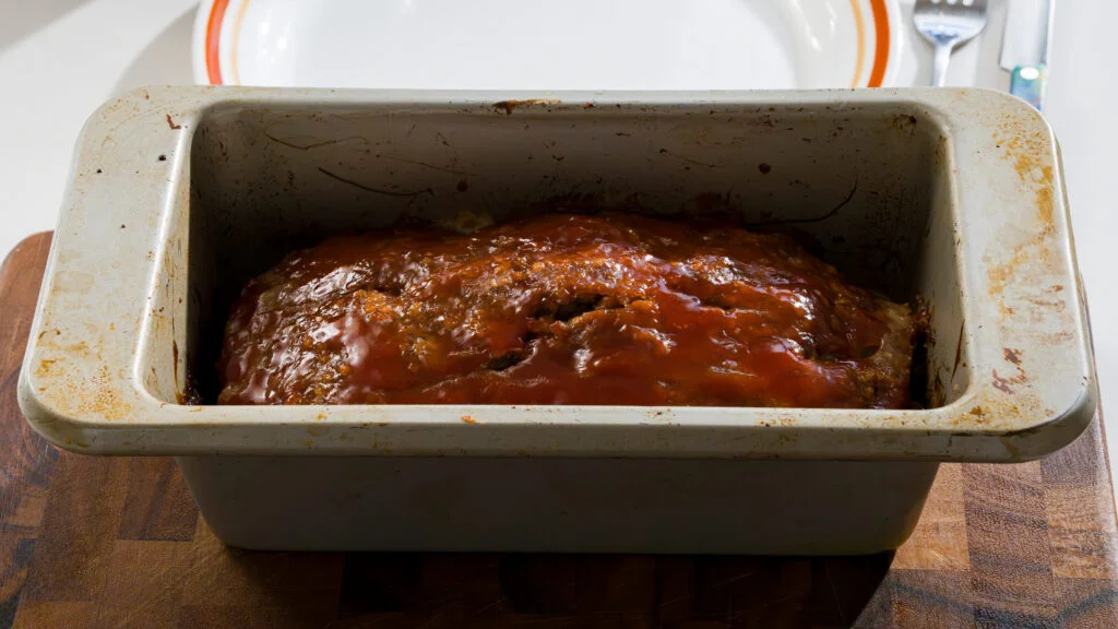A pair of hands brushing tangy ketchup glaze over a meatloaf on a parchment-lined sheet pan in a home kitchen