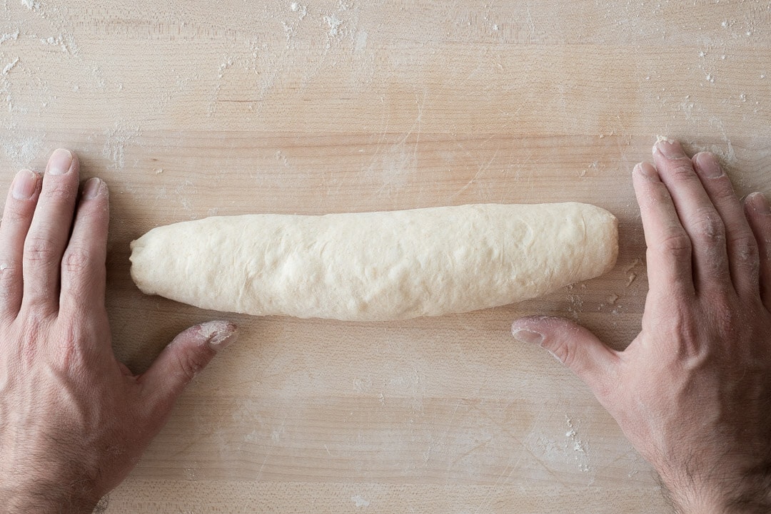 A pair of hands gently rolling and tapering baguette dough on a lightly floured wooden counter in a home kitchen