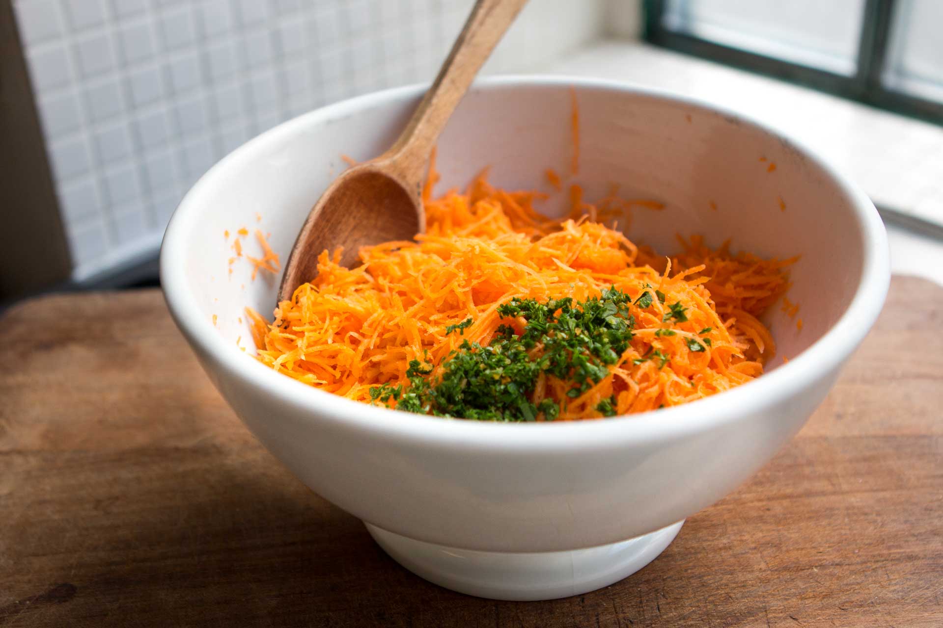 A pair of hands grating a carrot on a box grater over a large mixing bowl on a home kitchen counter