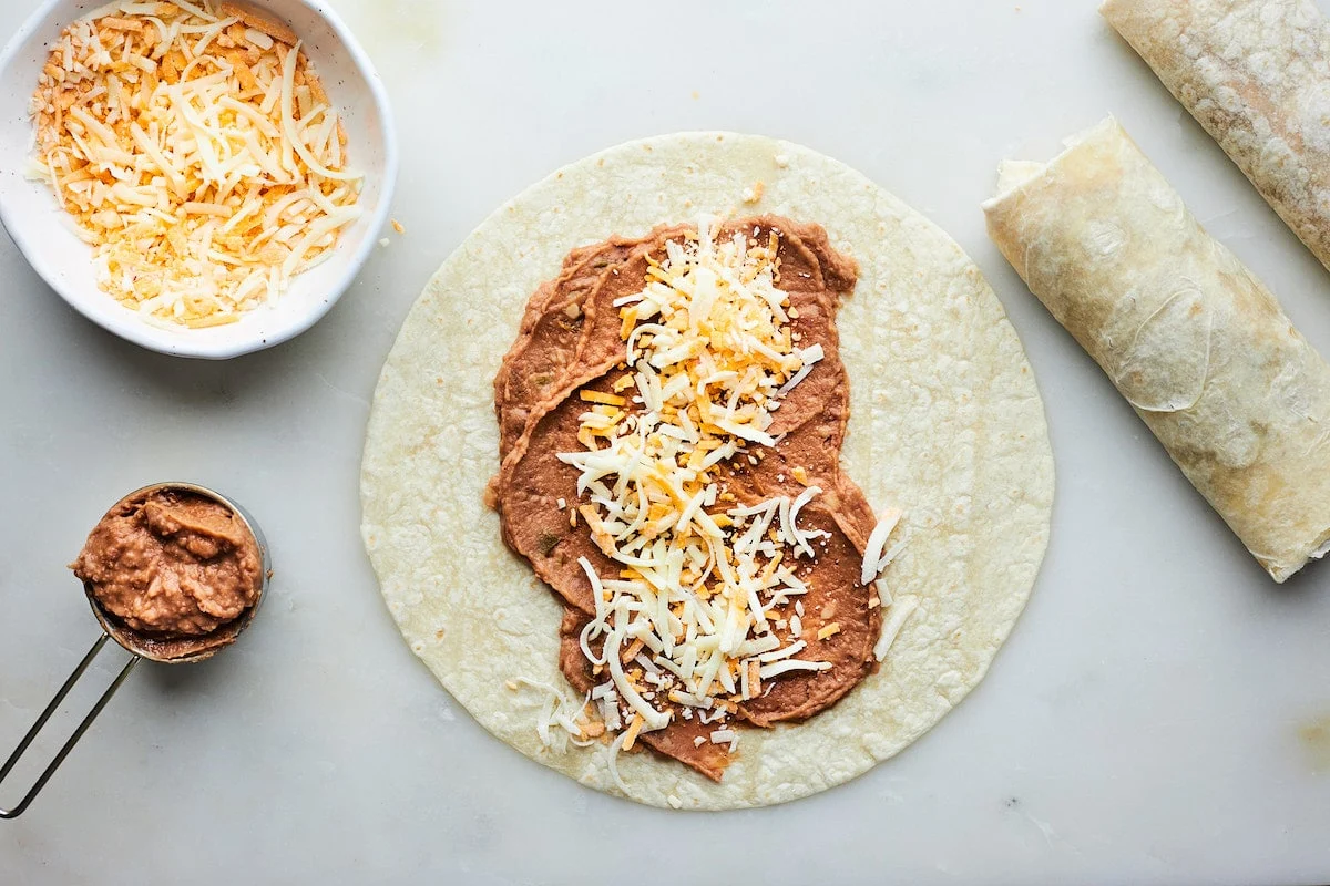 A pair of hands rolling a warm flour tortilla around a stripe of seasoned beans and shredded cheese on a wooden cutting board