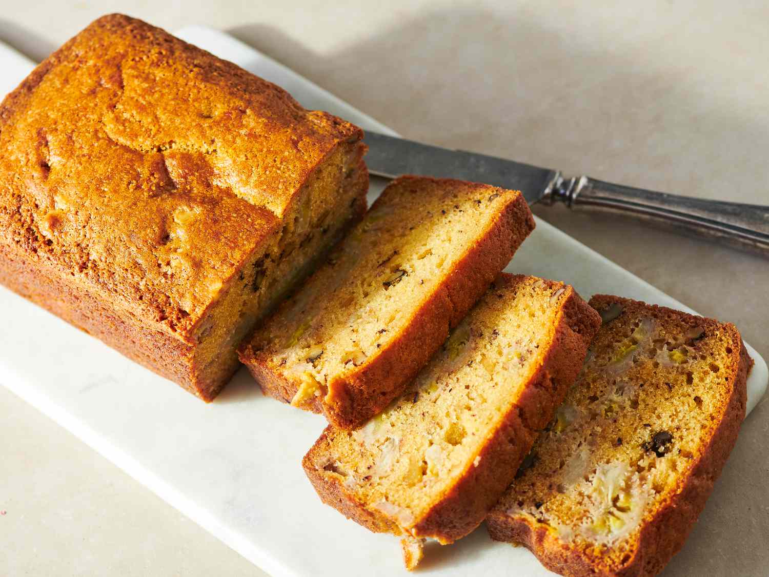 A pair of hands slicing a cooled loaf of banana bread with a serrated knife on a wooden cutting board in a home kitchen