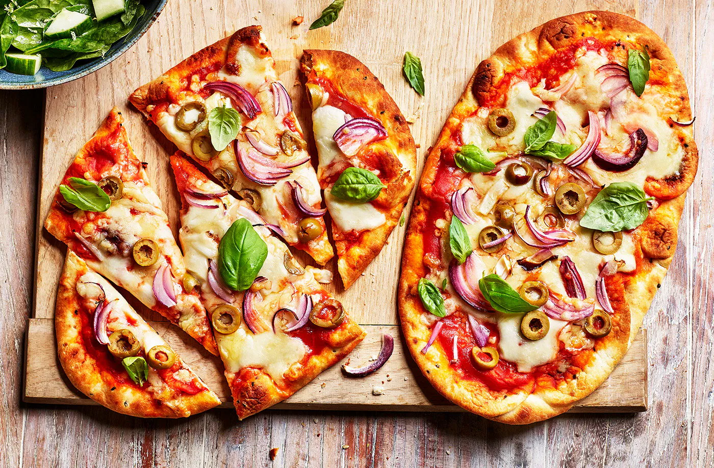 A pair of hands sprinkling shredded mozzarella and basil over sauced naan on a sheet pan