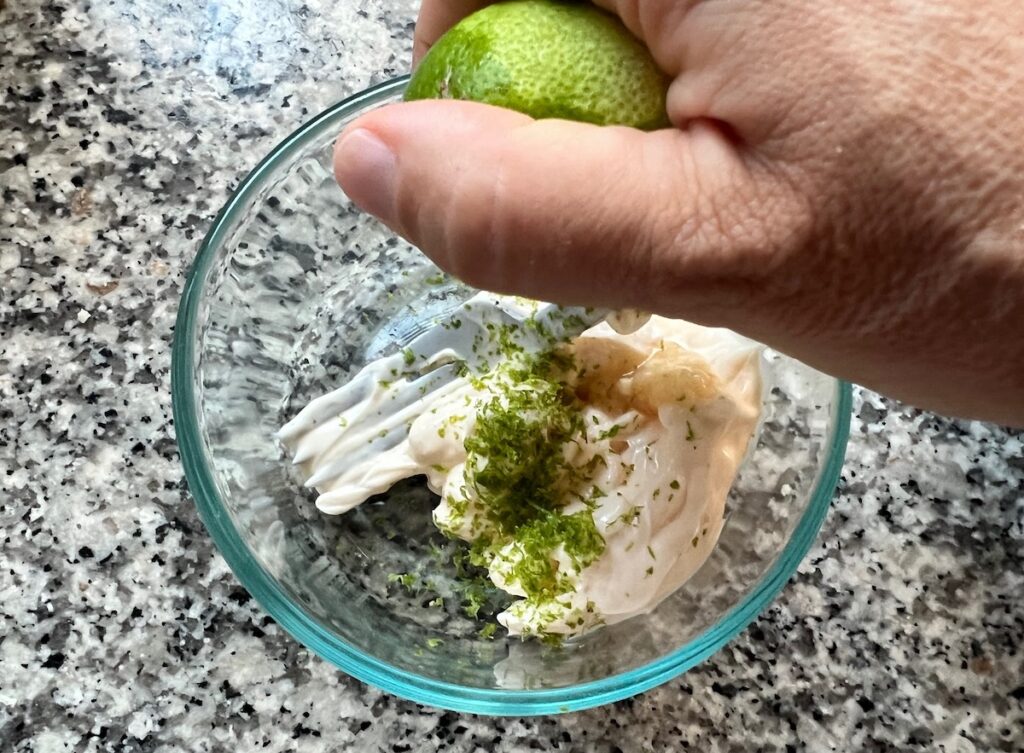 A pair of hands squeezing fresh lime juice over a bowl of black bean dip on a wooden cutting board in a cozy home kitchen