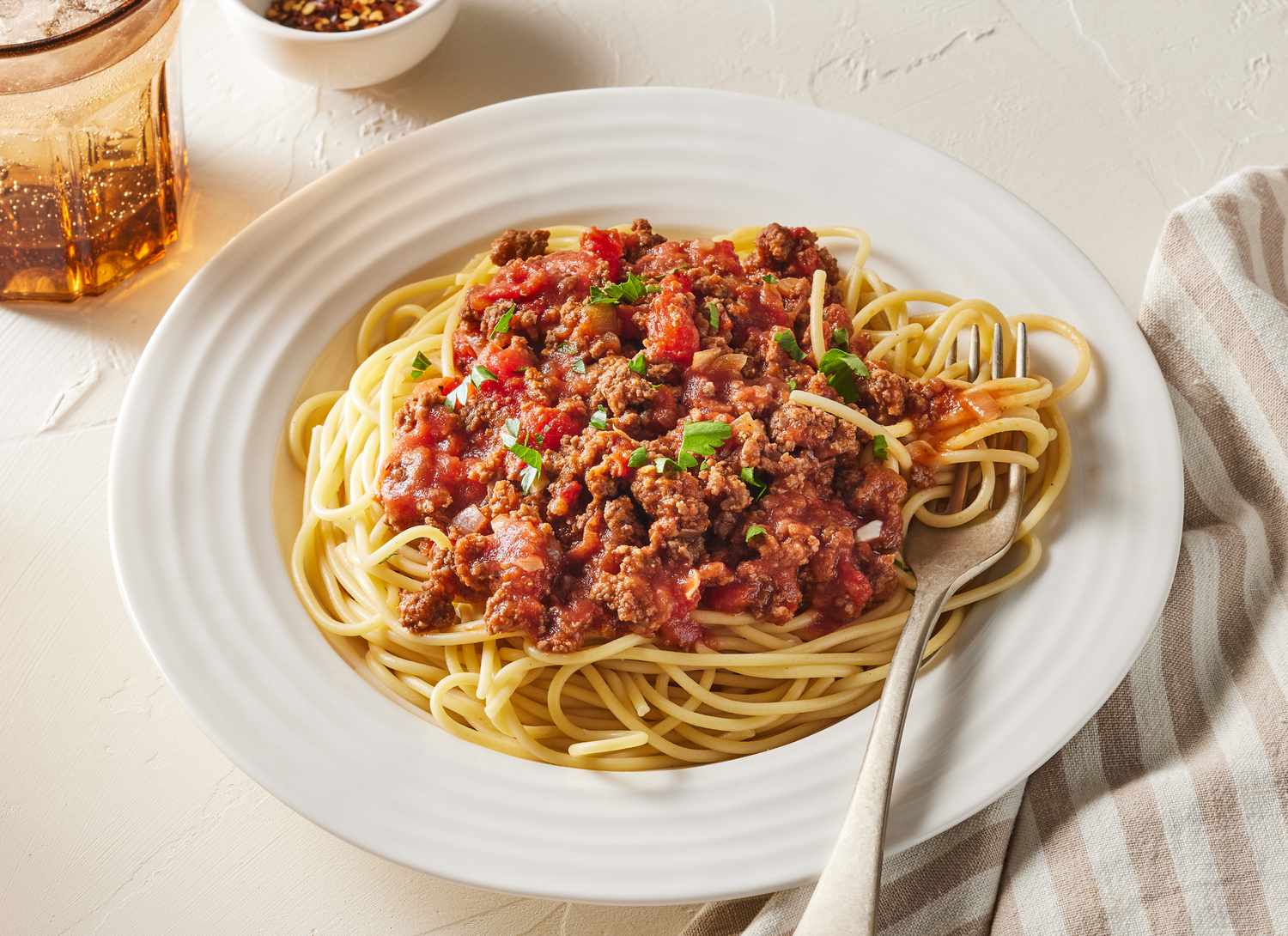 A pair of hands tossing cooked spaghetti with red meat sauce in a large stainless steel mixing bowl on a warm-lit kitchen counter