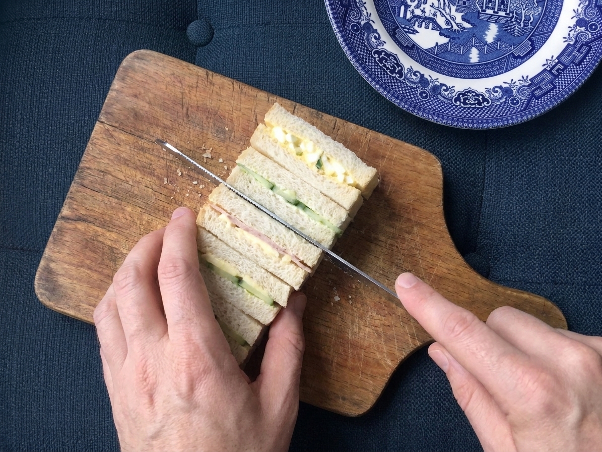 A pair of hands using a serrated knife to slice a chilled stack of finger sandwiches into neat rectangles on a wooden cutting board