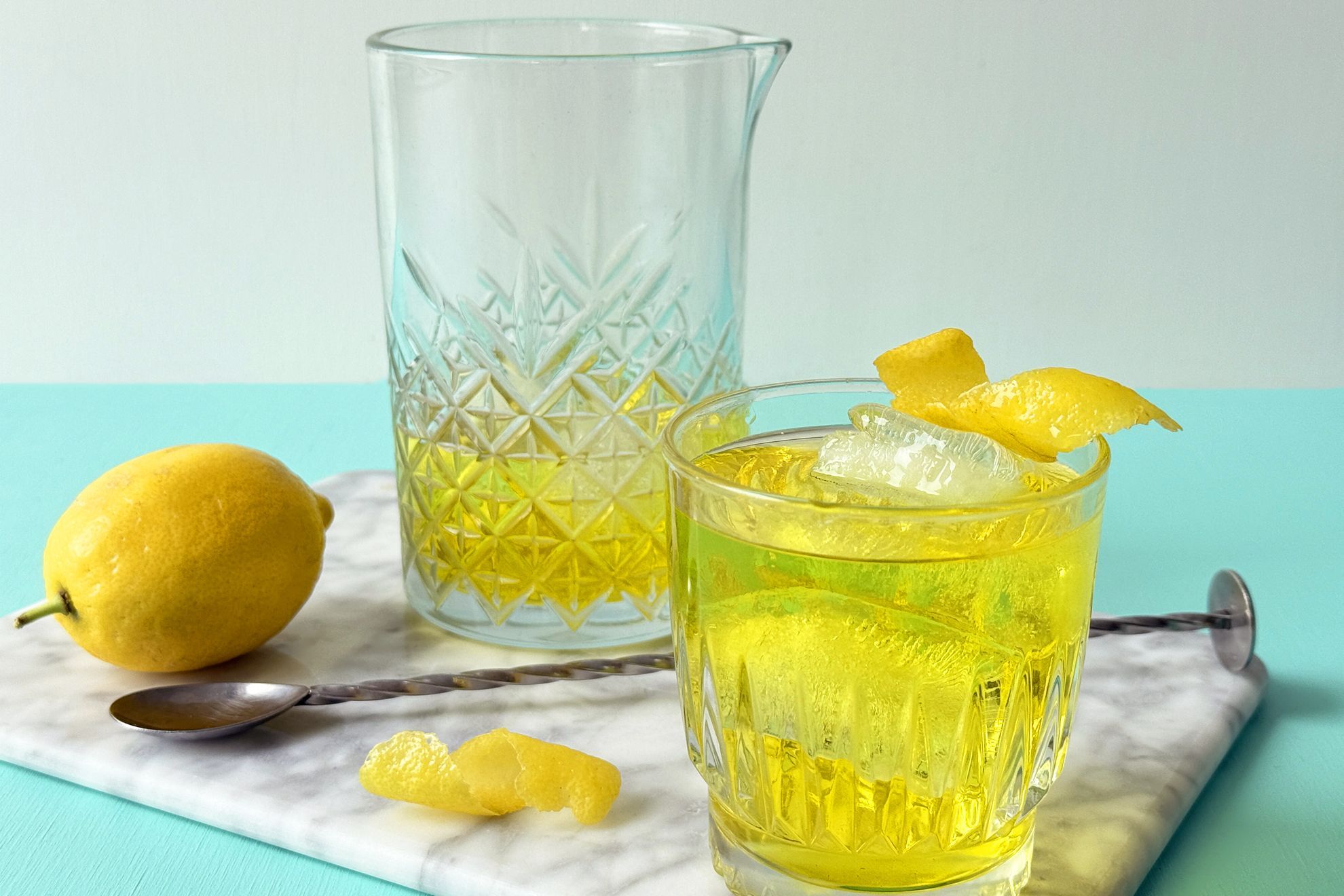 A pale golden White Negroni in a rocks glass over a large clear ice cube, garnished with a lemon twist on a warm kitchen counter with soft evening light