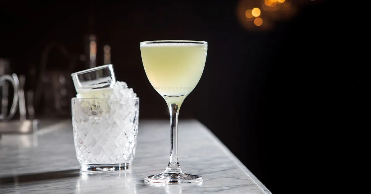 A pale green Last Word cocktail in a chilled coupe glass on a stone kitchen counter with a lime wheel nearby, warm home kitchen lighting