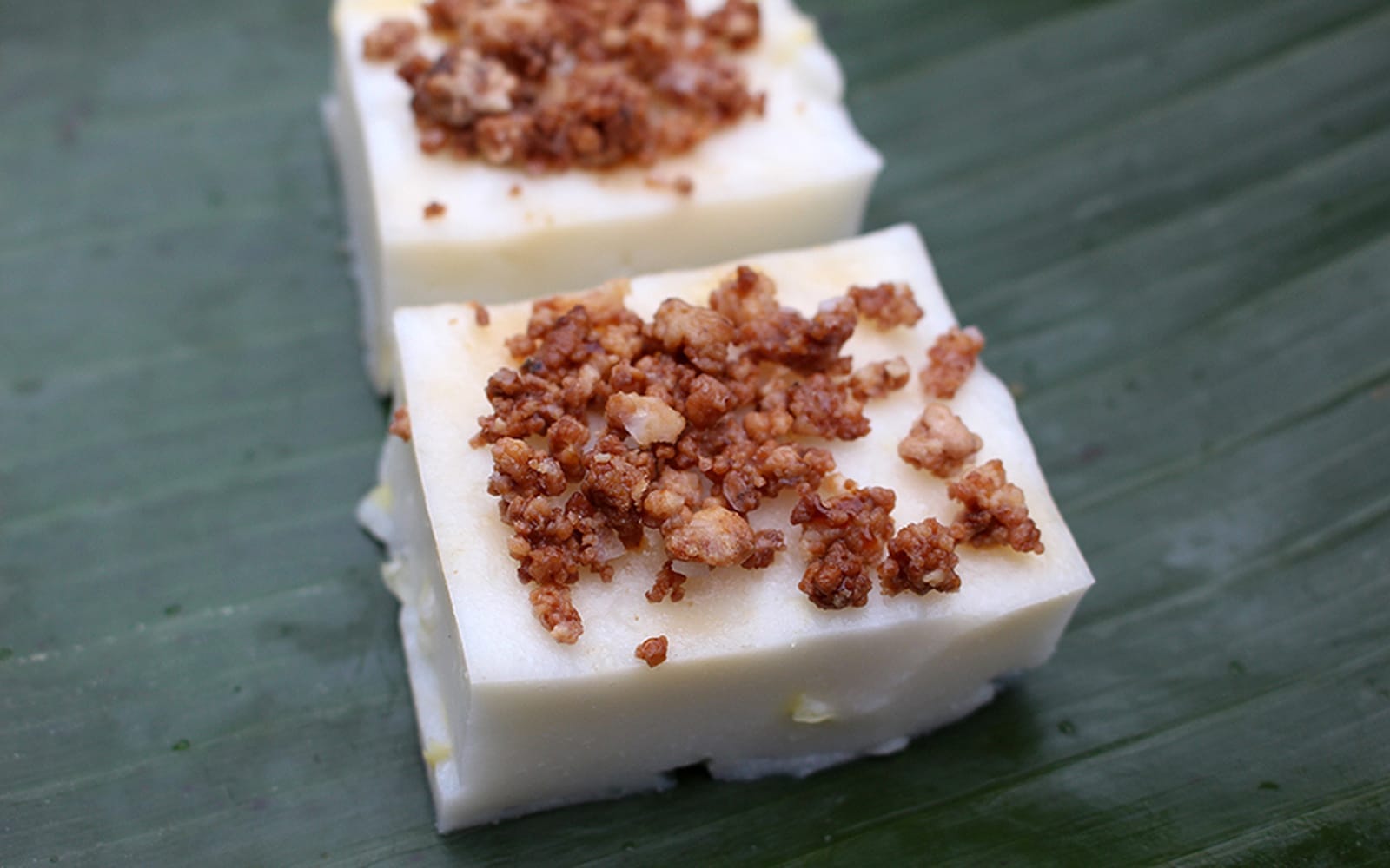 A pan of creamy maja blanca cut into neat squares, topped with golden toasted coconut latik on a simple wooden table with soft natural window light