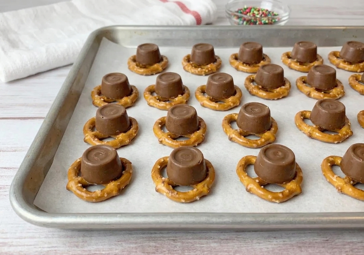 A parchment-lined baking sheet covered with mini pretzel snaps, each topped with an unwrapped Rolo candy, arranged in neat rows on a kitchen counter
