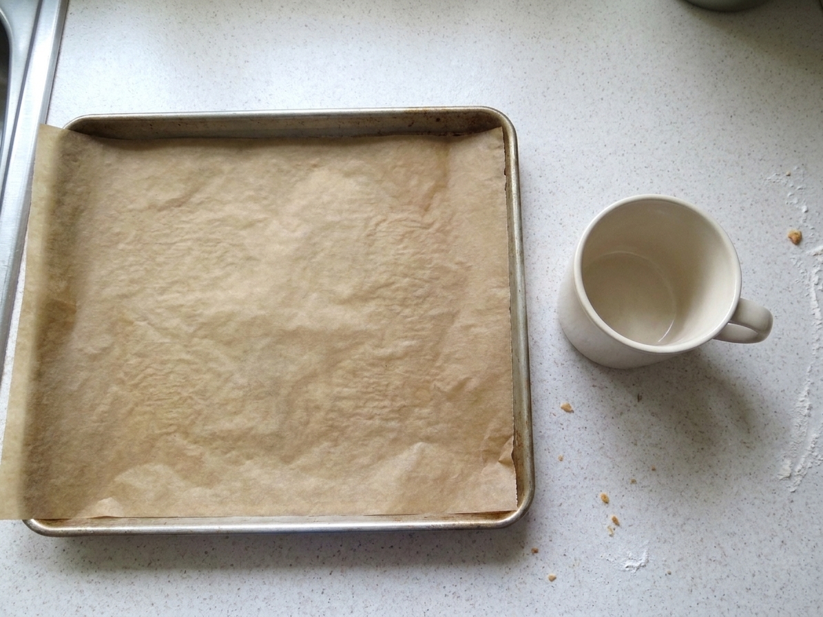 A parchment lined baking sheet on a kitchen counter next to a mug used for shaping fortune cookies