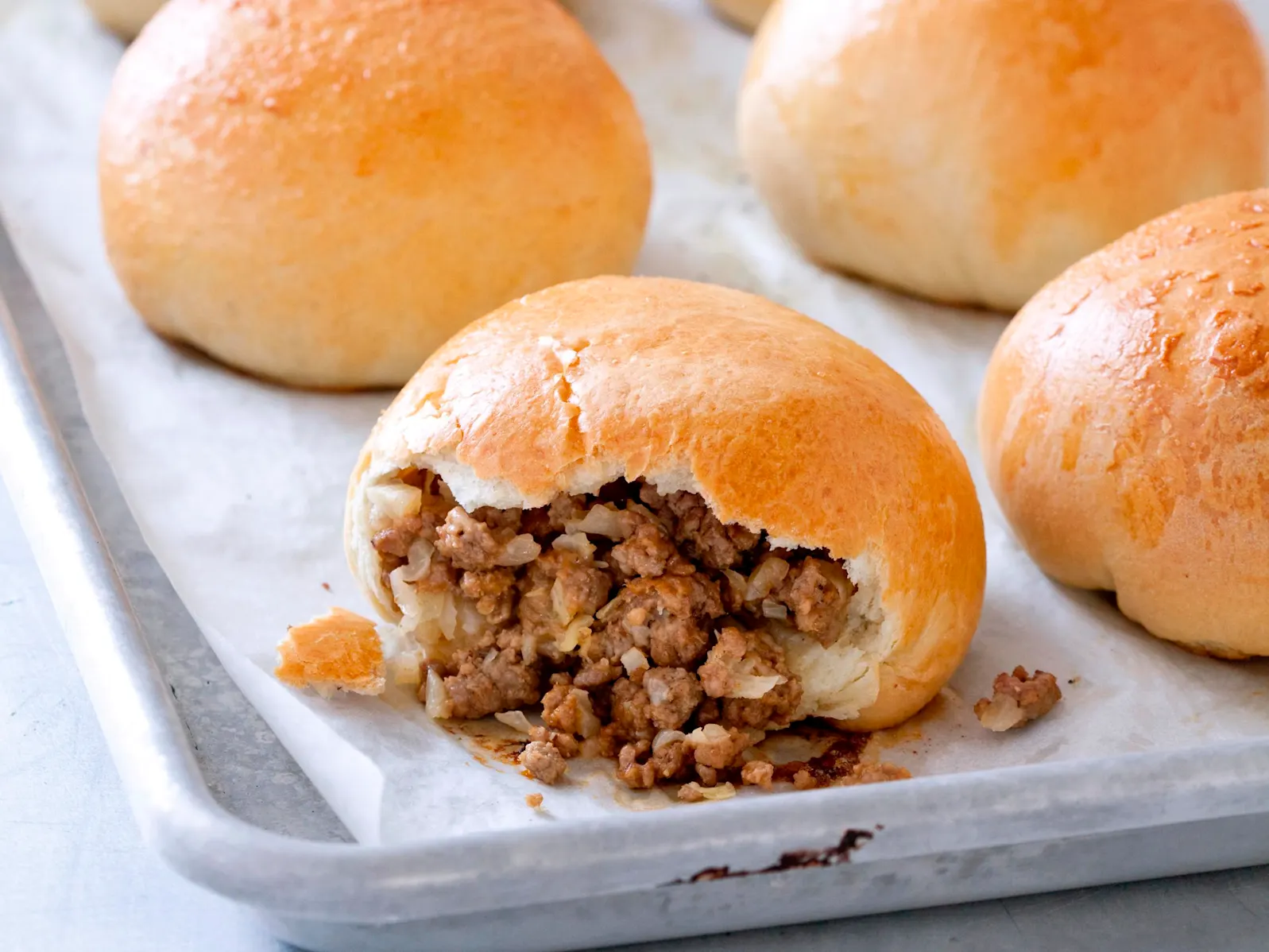 A parchment-lined sheet pan holding several golden-brown homemade Runza buns, one cut open to show the beef and cabbage filling, warm kitchen lighting